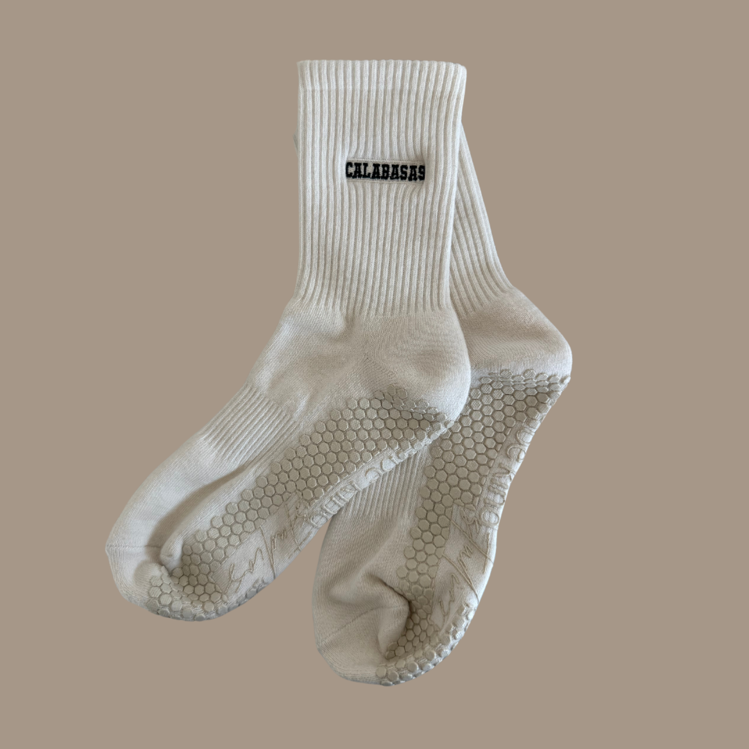 Beige grip socks with the word "CALABASAS" embroidered on the side horizontally & "be kind studios" embroidered on the bottom